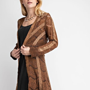 Pode incluir: Cardigã longo de renda marrom claro, aberto na frente, com detalhes em camurça e pequenos detalhes de tachas. O cardigã tem bainha e mangas com vieiras e é usado sobre um vestido preto. Um colar também é visível.