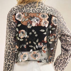 May include: A denim jacket with leopard print sleeves and back. The back has a sheer panel with floral embroidery and sequin embellishments. The model is wearing white shorts.