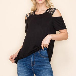 May include: Black off-the-shoulder top with rhinestone straps and short sleeves. The top is paired with blue denim jeans. The model is smiling and looking towards the camera.
