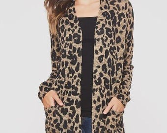 May include: A long, open-front cardigan with a leopard print pattern in shades of tan and black. The cardigan has long sleeves and two front pockets. The model is wearing a black top and blue jeans. The cardigan is a stylish outerwear option.