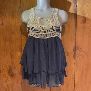 May include: A sleeveless top with a crochet bodice and a layered, flowing skirt. The bodice features a cream-colored crochet design with gold accents. The skirt is a dark navy blue, with multiple tiers of sheer fabric. The top is displayed on a mannequin.