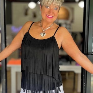 Puede incluir: Top negro con flecos, tirantes finos y diseño en capas. El top se combina con pantalones blancos y un collar con colgante de corazón plateado. La mujer sonríe frente a una puerta de cristal.