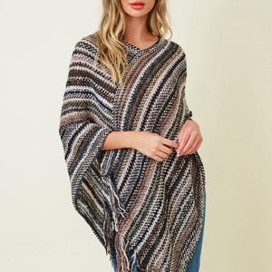 May include: A knit poncho featuring a multi-colored striped pattern in shades of black, white, brown, and grey. The garment has a V-neckline, a textured weave, and a fringed hemline along the bottom edge, providing a layered appearance.