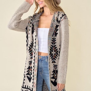 May include: A long, open-front cardigan in a neutral color with a Southwestern-inspired geometric pattern in black, orange, and teal. The cardigan is paired with a white top, blue jeans, and a beige hat. The sleeves are long and the cardigan reaches the mid-thigh.