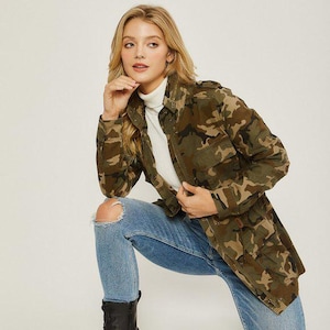 Puede incluir: Chaqueta con estampado de camuflaje en tonos verdes, marrones y beige. La chaqueta tiene múltiples bolsillos y una botonadura frontal. La modelo lleva un cuello alto blanco y vaqueros azules. Una opción de abrigo elegante.