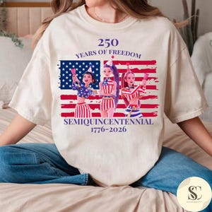 Puede incluir: Camiseta color crema con un diseño de la bandera estadounidense y tres figuras de dibujos animados. El texto dice "250 YEARS OF FREEDOM" y "SEMIQUINCENTENNIAL 1776-2026". El diseño utiliza los colores rojo, blanco y azul.