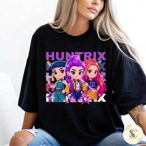 Puede incluir: Camiseta negra con la palabra "HUNTRIX" sobre una imagen de dibujos animados de tres chicas con pelo y atuendos coloridos. Las chicas sostienen varitas en forma de estrella. El diseño incluye estrellas moradas y un fondo blanco.