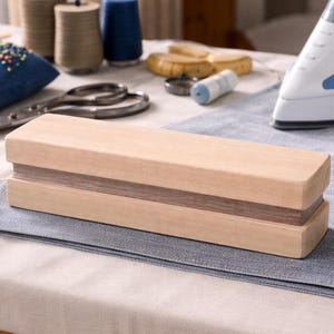 May include: A wooden clapper, a rectangular tool with a light wood exterior and a darker wood interior, rests on a gray fabric. Sewing supplies, including spools of thread, a blue pin cushion, and scissors, are in the background.