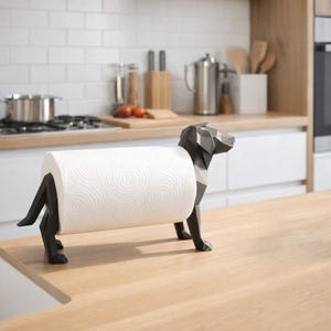 Könnte beinhalten: Ein schwarzer, geometrischer Papiertuchhalter in Form eines Hundes. Der Halter hält eine Rolle weißer Papiertücher. Der Hund steht auf einer hellen Holz-Arbeitsplatte in einer Küche, mit einer gefliesten Rückwand.