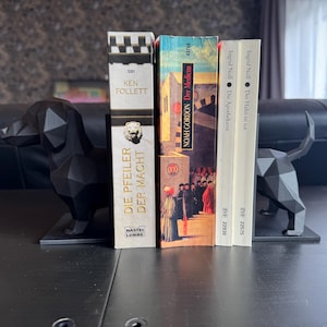 Può includere: Un paio di fermalibri neri a forma di cane geometrico che sostengono diversi libri. I libri includono titoli come "Die Pfeiler der Macht" e "Der Medicus". I fermalibri sono realizzati in un materiale scuro e hanno un design moderno e angolare.