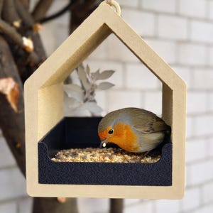 May include: A beige, house-shaped bird feeder with a black feeding tray, suspended by a natural fiber cord. A small bird with orange and brown plumage is perched inside, eating seeds. The feeder is hanging in front of a white brick wall.