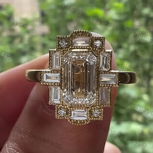 Op de afbeelding: Een gouden ring met een smaragdgeslepen middensteen en baguette-geslepen zijstenen. De ring heeft een geometrisch ontwerp en is bezet met kleine accentstenen. De ring wordt door een persoon vastgehouden.