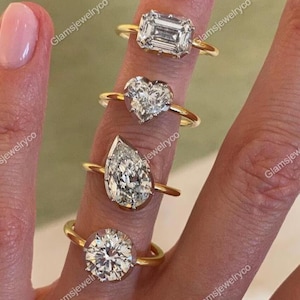 Könnte beinhalten: Eine Hand zeigt vier goldene Ringe mit Diamanten in verschiedenen Schliffen: Smaragd, Herz, Tropfen und rund. Die Ringe sind auf einem goldenen Band gefasst. Der Hintergrund ist hellbeige.