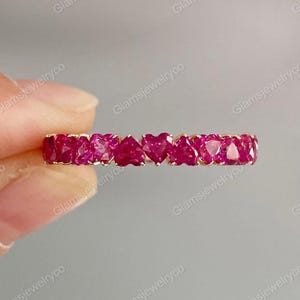 Puede incluir: Un anillo de oro con una serie de piedras preciosas rojas vibrantes en forma de corazón. Las piedras están engastadas muy juntas, creando una banda continua de color. El anillo se muestra sobre un fondo neutro.