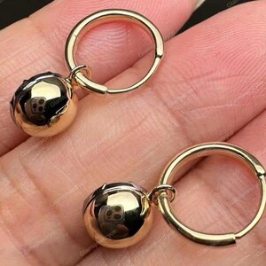 May include: Two gold-colored hoop earrings with a spherical charm. The earrings are made of a shiny, reflective metal. The spherical charm has a textured surface. The earrings are shown against a skin-toned background.