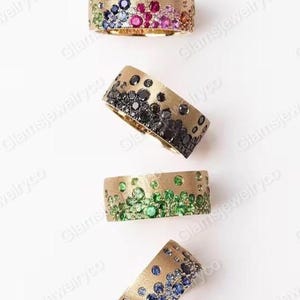 May include: Four gold rings with a textured finish, each set with different gemstones. One ring has a rainbow of colours, another has black stones, a third has green stones, and the last has blue stones.