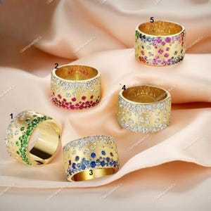 Op de afbeelding: Vijf gouden ringen met kleurrijke edelsteenaccenten worden gepresenteerd op een perzikroze stof. De ringen hebben verschillende edelsteenkleuren, waaronder groen, rood, blauw en een regenboog aan tinten. Elke ring heeft een uniek ontwerp.
