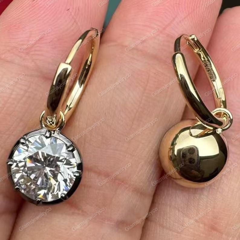 May include: A pair of gold-colored hoop earrings. One earring features a large, round, clear gemstone set in a dark metal bezel. The other earring has a smooth, gold-colored ball. The earrings are shown against a skin-toned background.