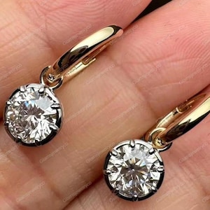 May include: A pair of gold hoop earrings with a large, round, sparkling diamond pendant. The diamonds are set in a dark metal bezel, and the hoops are a polished gold color. The earrings are shown against a skin-toned background.