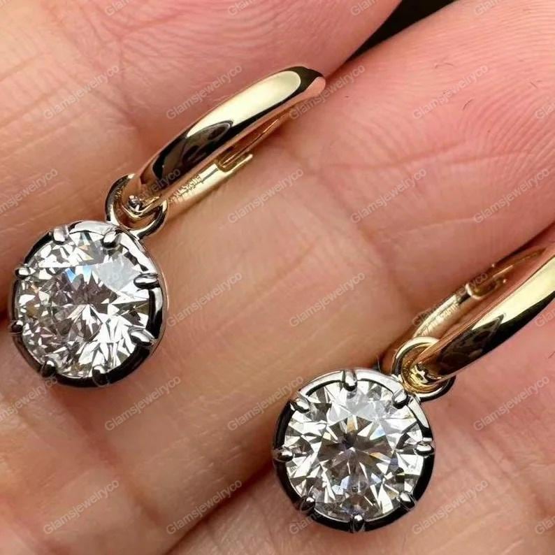 May include: A pair of gold hoop earrings with a large, round, sparkling diamond pendant. The diamonds are set in a dark metal bezel, and the hoops are a polished gold color. The earrings are shown against a skin-toned background.