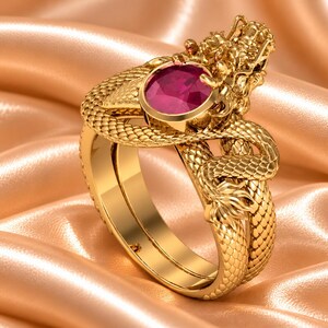 Puede incluir: Un anillo de tono dorado con un diseño de dragón esculpido que se envuelve alrededor de la banda. El cuerpo del dragón tiene escamas detalladas y la cabeza está colocada sobre una piedra preciosa ovalada de color rojo intenso en un engaste liso.