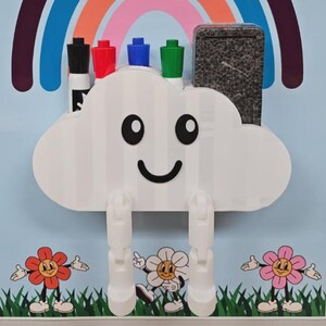 May include: A white cloud-shaped wall organizer with a smiling face, two dangling white legs, and slots holding four markers and a black eraser. The organizer is mounted on a rectangular board featuring a rainbow, green grass, and three flowers.