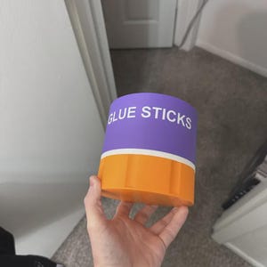 May include: A cylindrical storage container with a purple top section featuring the white text GLUE STICKS and a solid orange bottom section. The container has a smooth finish and a distinct horizontal white band separating the purple and orange parts.