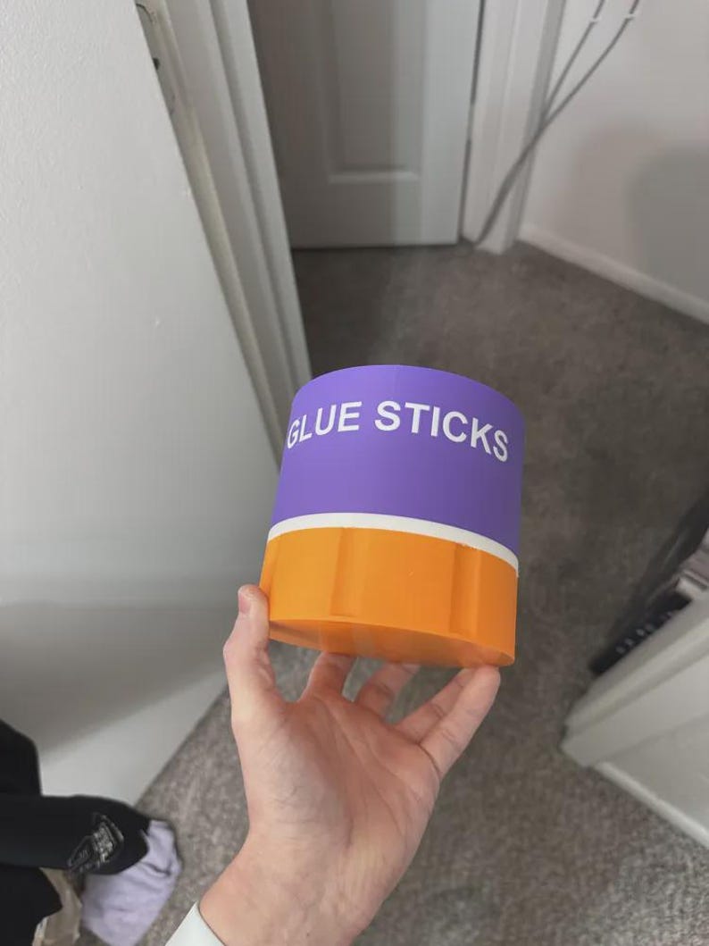 May include: A cylindrical storage container with a purple top section featuring the white text GLUE STICKS and a solid orange bottom section. The container has a smooth finish and a distinct horizontal white band separating the purple and orange parts.