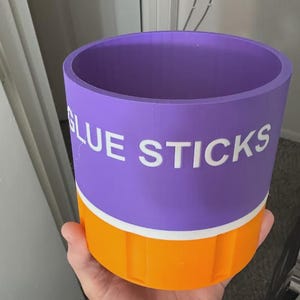 May include: A cylindrical purple and orange container with a white horizontal stripe separating the two colors. The word GLUE STICKS is printed in white capital letters on the purple section. The container has a smooth finish and a hollow interior.