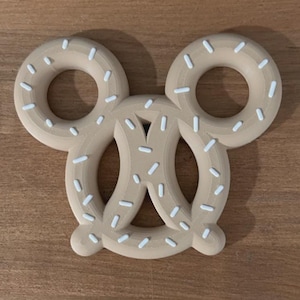 Puede incluir: Un mordedor de silicona de color beige con forma de pretzel, que presenta dos bucles circulares superiores y una sección central anudada. La superficie está decorada con pequeñas chispas blancas en forma de guion distribuidas por todo el objeto.