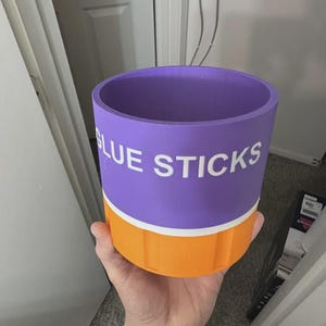 May include: A cylindrical purple and orange container with a white horizontal stripe separating the two colors. The text GLUE STICKS is printed in white capital letters on the purple section. The container has a smooth top rim and vertical ridges.