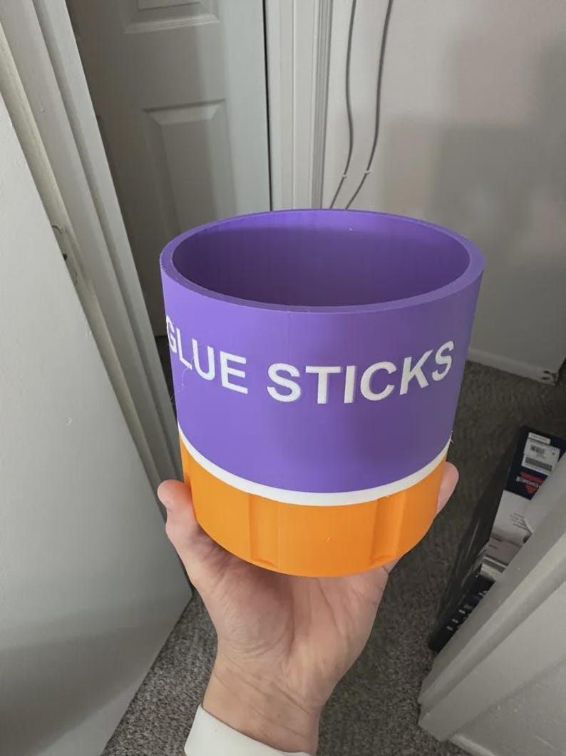May include: A cylindrical purple and orange container with a white horizontal stripe separating the two colors. The text GLUE STICKS is printed in white capital letters on the purple section. The container has a smooth top rim and vertical ridges.