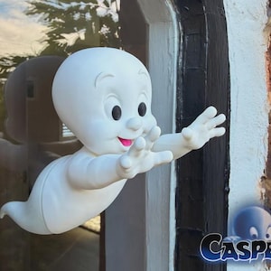 Könnte beinhalten: Eine weiße Casper-Figur mit rundem Kopf, großen schwarzen Augen und einem lächelnden Mund mit rosa Zunge. Die Figur hat ausgestreckte Arme und Hände, die nach vorne greifen und so aussehen, als würden sie aus einer Glasoberfläche hervorkommen.