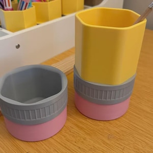 May include: Two hexagonal pencil holders designed to resemble pencils. One is a short, wide container with a pink base, a grey ribbed band, and a grey top. The other is a taller, upright container with a yellow top, a grey ribbed band, and a pink base.