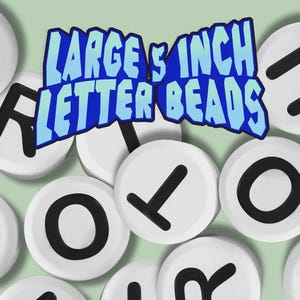 Pode incluir: Contas de letras redondas brancas com letras pretas. A imagem inclui o texto "LARGE 12,7 CM LETTER BEADS" em uma fonte azul em estilo cartoon. As contas estão espalhadas em um fundo verde claro.