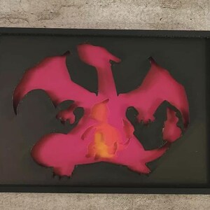 May include: A rectangular black shadow box frame containing a layered paper cutout of a dragon-like creature. The silhouette is primarily deep pink, with a bright yellow and orange gradient glowing from the center of the creature's body.