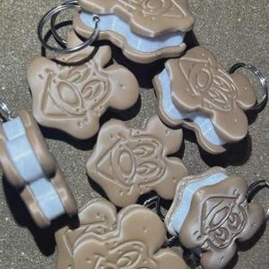 May include: A collection of light brown, cookie-shaped keychains featuring a debossed cartoon face design. Each keychain consists of two brown layers with a light blue center layer and includes a silver metal split ring for attachment.