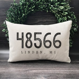 May include: A white pillow with black text that reads "48566 Linden, MI". The pillow is sitting on a wooden surface.