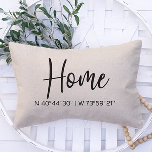 May include: A beige linen pillow with the word "Home" in black cursive script and the coordinates "N 40°44' 30" | W 73°59' 21" printed below.