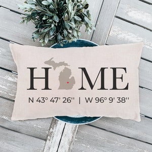 May include: A beige throw pillow with the word "HOME" in black text and an outline of the state of Michigan with a red heart in the center. The coordinates "N 43° 47' 26" | W 96° 9' 38" are printed below the word "HOME".