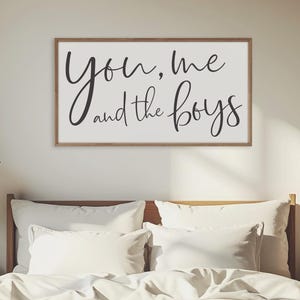 May include: A white wooden sign with black calligraphy that reads "You, Me and the Boys". The sign is hanging on a wall above a white bed with a wooden headboard.