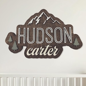 May include: Wooden wall art featuring a mountain silhouette with the names "HUDSON" and "carter" in white lettering. The dark brown sign includes two green pine trees and white mountain peaks. The sign is mounted above a white crib.