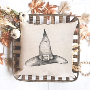 May include: A beige pillow with a black and white illustration of a witch's hat. The hat has a wide brim and a tall pointed crown with a buckle.