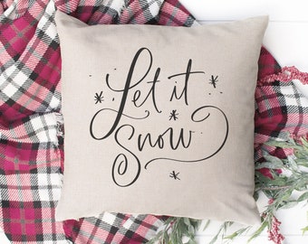 May include: A beige throw pillow with the text "Let it snow" written in black cursive script. The pillow is surrounded by a red and white plaid blanket and green foliage.