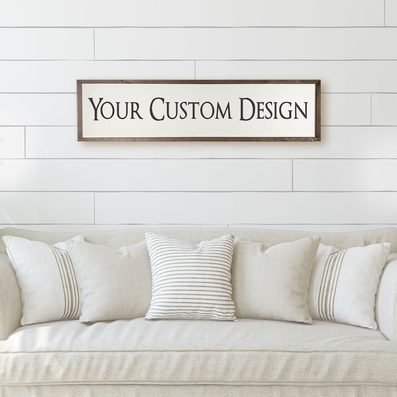 Puede incluir: Un letrero de madera blanco con marco marr&oacute;n, colgado en una pared blanca. El letrero dice "YOUR CUSTOM DESIGN". El letrero est&aacute; sobre un sof&aacute; beige con almohadas de rayas blancas y marrones.