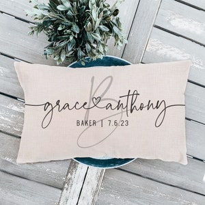 May include: A beige pillow with the text "grace & anthony BAKER | 7.6.23" in black cursive font. The text is centered on the pillow and is surrounded by a decorative gray heart and a stylized "B".