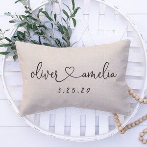 May include: A beige linen pillow with the names "oliver" and "amelia" written in cursive script with a heart symbol between them. The date "3.25.20" is written below the names.