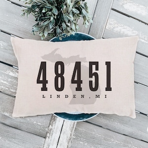 May include: A beige throw pillow with the number 48451 and the text "Linden, MI" printed in black. The pillow features a silhouette of the state of Michigan.