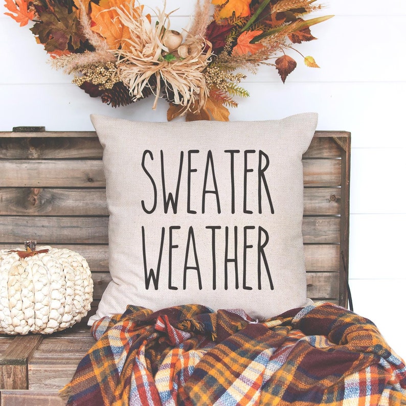 May include: A white throw pillow with black text that reads "sweater weather" on a wooden bench with a plaid throw blanket and a white pumpkin.