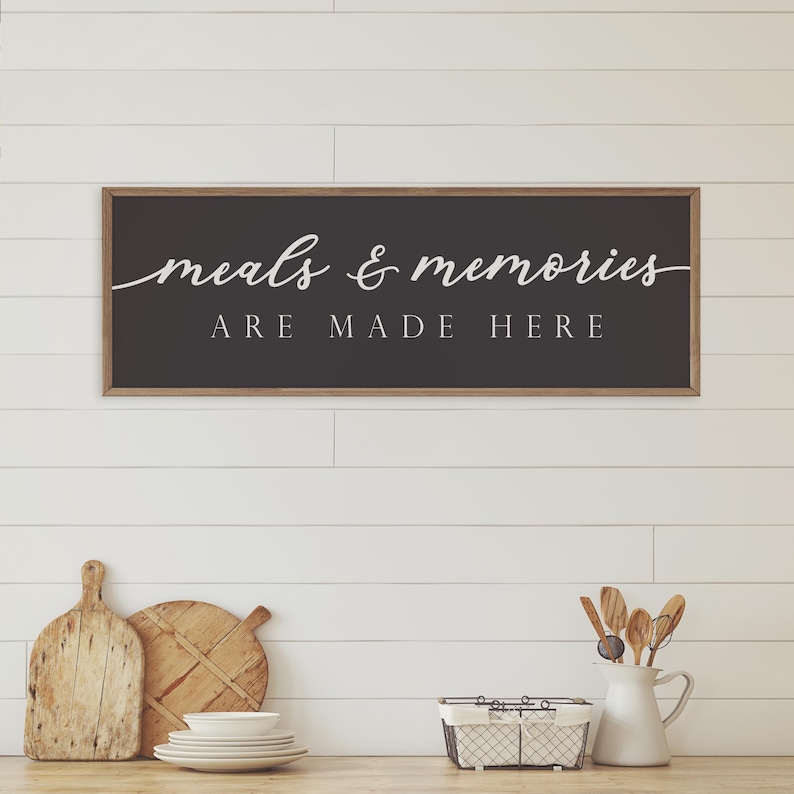 May include: A black framed sign with white text that reads "meals & memories are made here" hanging on a white shiplap wall.